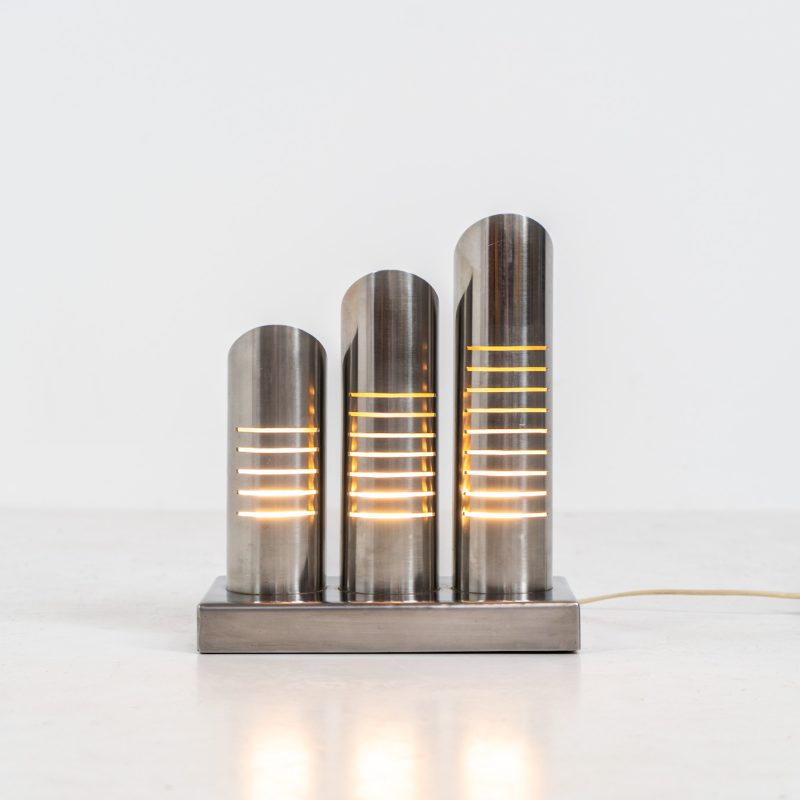 Space age table lamp by Goffredo Reggiani (Italy, 1970s). This unique piece is made out of brushed stainless steel and consists of three cylindrical tubes with milled slits which creates a stunning light glow. The lamp seamlessly blends form and function and is a true statement piece. It is in very good condition with some patina on it.