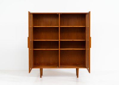 Teak showcase by Ib Kofod-Larsen for Faarup Møbelfabrik (Denmark, 1960s). This beautiful showcase features a stunning teak woodgrain with intricate details throughout. It has two doors with elegant handles and glass panels, providing a clear view of the interior. Inside, there are three adjustable shelves, offering lots of storage options. It is in perfect condition and labeled by maker.
