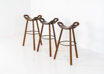 Set of 3 Brutalist “Marbella” stools manufactured by Conoform and attributed to Sergio Rodrigues (Spain, 1970s). This iconic set of Marbella stools, also known as the “Spanish Stool,” exemplifies brutalist design with a sculptural flair. Crafted from solid beech wood, the stools feature a distinctive curved seat and are complemented by a sturdy iron footrest. The set is in good vintage condition, displaying some signs of use such as seat marks and wear consistent with age.