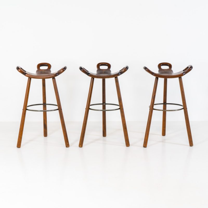 Set of 3 Brutalist “Marbella” stools manufactured by Conoform and attributed to Sergio Rodrigues (Spain, 1970s). This iconic set of Marbella stools, also known as the “Spanish Stool,” exemplifies brutalist design with a sculptural flair. Crafted from solid beech wood, the stools feature a distinctive curved seat and are complemented by a sturdy iron footrest. The set is in good vintage condition, displaying some signs of use such as seat marks and wear consistent with age.