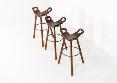 Set of 3 Brutalist “Marbella” stools manufactured by Conoform and attributed to Sergio Rodrigues (Spain, 1970s). This iconic set of Marbella stools, also known as the “Spanish Stool,” exemplifies brutalist design with a sculptural flair. Crafted from solid beech wood, the stools feature a distinctive curved seat and are complemented by a sturdy iron footrest. The set is in good vintage condition, displaying some signs of use such as seat marks and wear consistent with age.