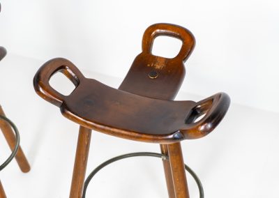 Set of 3 Brutalist “Marbella” stools manufactured by Conoform and attributed to Sergio Rodrigues (Spain, 1970s). This iconic set of Marbella stools, also known as the “Spanish Stool,” exemplifies brutalist design with a sculptural flair. Crafted from solid beech wood, the stools feature a distinctive curved seat and are complemented by a sturdy iron footrest. The set is in good vintage condition, displaying some signs of use such as seat marks and wear consistent with age.