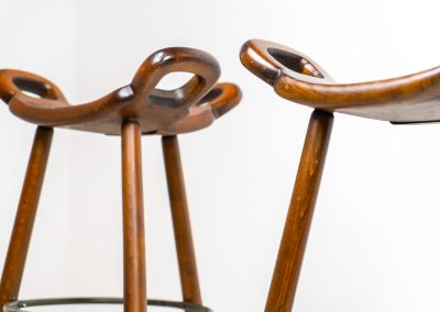 Set of 3 Brutalist “Marbella” stools manufactured by Conoform and attributed to Sergio Rodrigues (Spain, 1970s). This iconic set of Marbella stools, also known as the “Spanish Stool,” exemplifies brutalist design with a sculptural flair. Crafted from solid beech wood, the stools feature a distinctive curved seat and are complemented by a sturdy iron footrest. The set is in good vintage condition, displaying some signs of use such as seat marks and wear consistent with age.