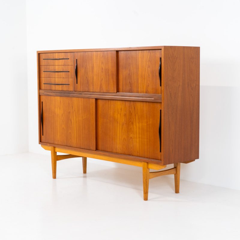 Danish highboard in teak, 1960s. A beautiful mid-century piece crafted in rich teak and standing on oak base, featuring lots of storage space and contrasting black handels. The highboard showcases a warm, natural woodgrain, sleek lines, and distinctive handles. It is in excellent condition with some minor using traces on the top.