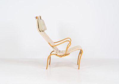 “Miranda” easy chair by Bruno Mathsson Design Studio (Sweden, 1970s). Originally designed by Bruno Mathsson in 1941, this exceptional “Miranda” easy chair was manufactured in the 1970s. It features a beautifully curved, molded beech plywood frame with a linen webbing seat and an adjustable headrest, suspended by leather straps. The chair is in good vintage condition, showing signs of use such as marks, fabric fraying, and some damage to the wood, all of which add to its authentic character. Marked by maker.