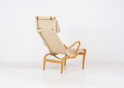 “Miranda” easy chair by Bruno Mathsson Design Studio (Sweden, 1970s). Originally designed by Bruno Mathsson in 1941, this exceptional “Miranda” easy chair was manufactured in the 1970s. It features a beautifully curved, molded beech plywood frame with a linen webbing seat and an adjustable headrest, suspended by leather straps. The chair is in good vintage condition, showing signs of use such as marks, fabric fraying, and some damage to the wood, all of which add to its authentic character. Marked by maker.