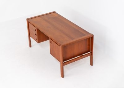 Rare executive writing desk by H.P. Hansen (Denmark, 1950s). This large and exceptionally beautiful piece is a true example of top-quality Danish craftsmanship. Made from solid teak, it showcases elegant aesthetics with softly rounded edges and sculptural inset handles. The spacious desktop highlights a striking wood grain and is complemented by three drawers, a cabinet, and two open shelves at the back. The desk is finished on both sides, making it an ideal statement piece for placement in the center of a room. It is in excellent condition, with only minor discoloration on the top and light signs of use on the drawers.