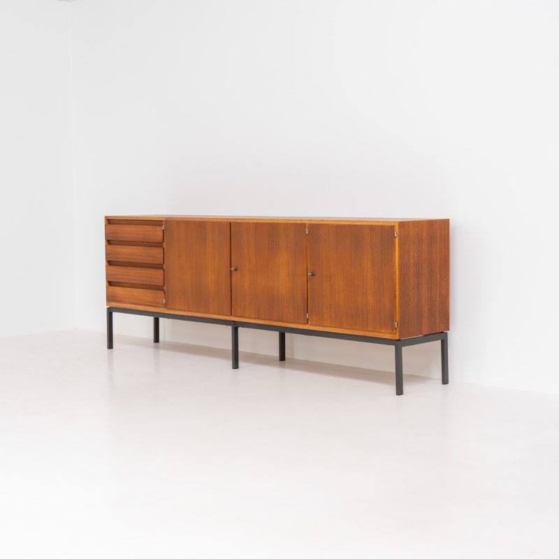 XL Modernist sideboard by Behr Möbel (Germany, 1950s). This striking modernist sideboard was produced in the 1950s by the renowned German manufacturer Behr Möbel, best known for the iconic B40 model. Expertly crafted from rich teak wood, the piece rests on a sleek metal base that adds to its architectural presence. It features four drawers and three doors, offering generous storage space with adjustable shelves inside. The sideboard is in very good condition, with fully functional locks.