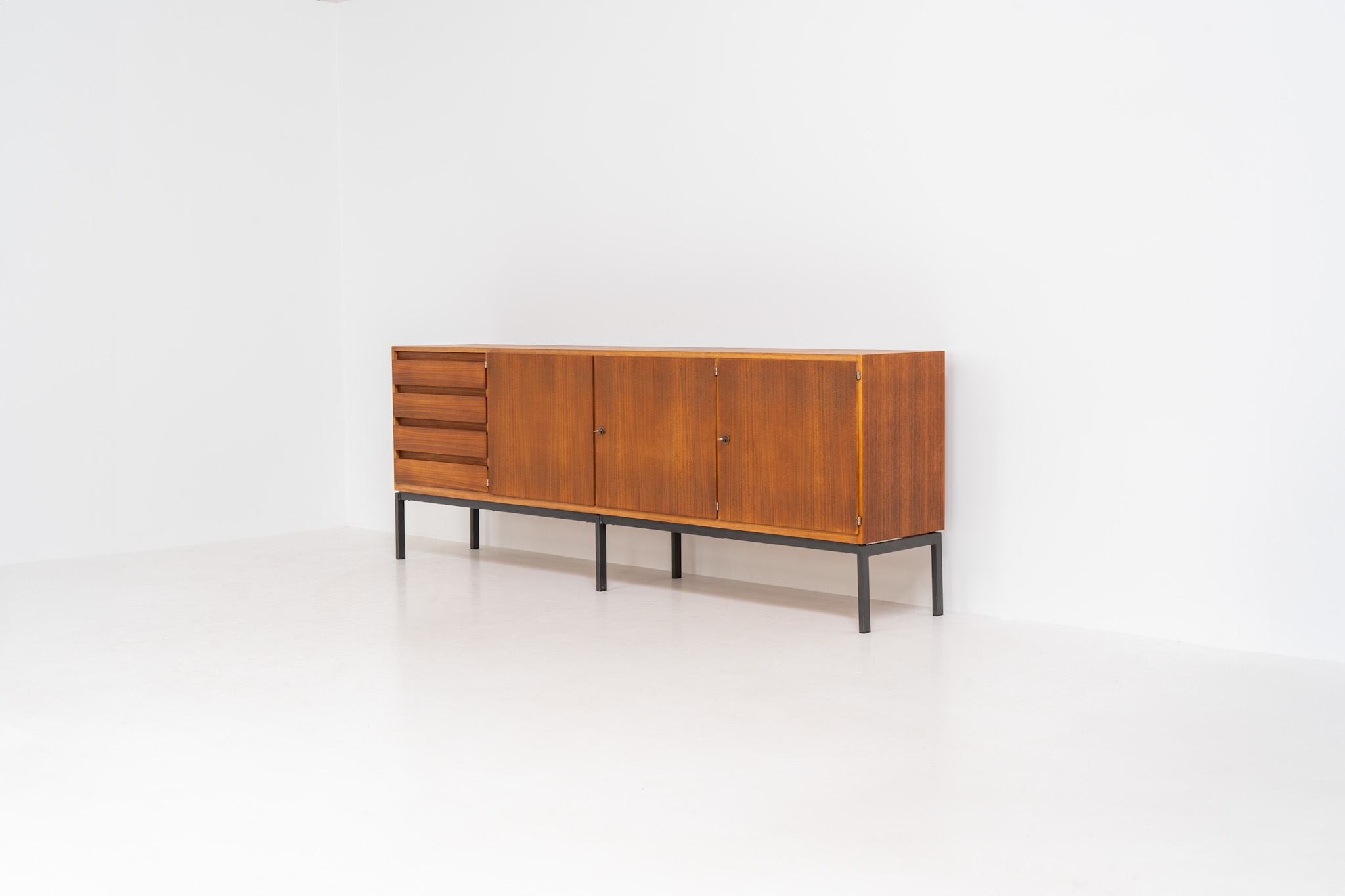 XL Modernist sideboard by Behr Möbel (Germany, 1950s). This striking modernist sideboard was produced in the 1950s by the renowned German manufacturer Behr Möbel, best known for the iconic B40 model. Expertly crafted from rich teak wood, the piece rests on a sleek metal base that adds to its architectural presence. It features four drawers and three doors, offering generous storage space with adjustable shelves inside. The sideboard is in very good condition, with fully functional locks.