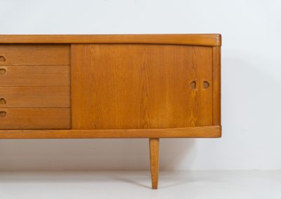 Exceptional sideboard by Henry W. Klein for Bramin in oak (Denmark, 1960s). This piece is a true masterpiece of Danish mid-century design. Crafted from high-quality materials, it showcases exquisite attention to detail, from the sculptural integrated handles to the beautifully crafted corner joints. The sideboard offers the perfect balance of aesthetics and functionality. It features two large sliding doors with adjustable shelves behind them and four drawers. The highboard is in very good condition and marked by maker.