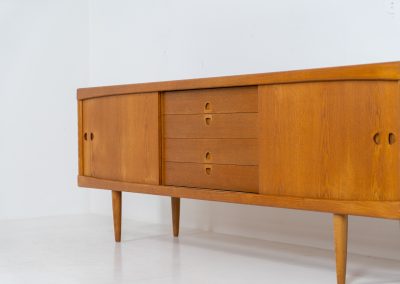 Exceptional sideboard by Henry W. Klein for Bramin in oak (Denmark, 1960s). This piece is a true masterpiece of Danish mid-century design. Crafted from high-quality materials, it showcases exquisite attention to detail, from the sculptural integrated handles to the beautifully crafted corner joints. The sideboard offers the perfect balance of aesthetics and functionality. It features two large sliding doors with adjustable shelves behind them and four drawers. The highboard is in very good condition and marked by maker.
