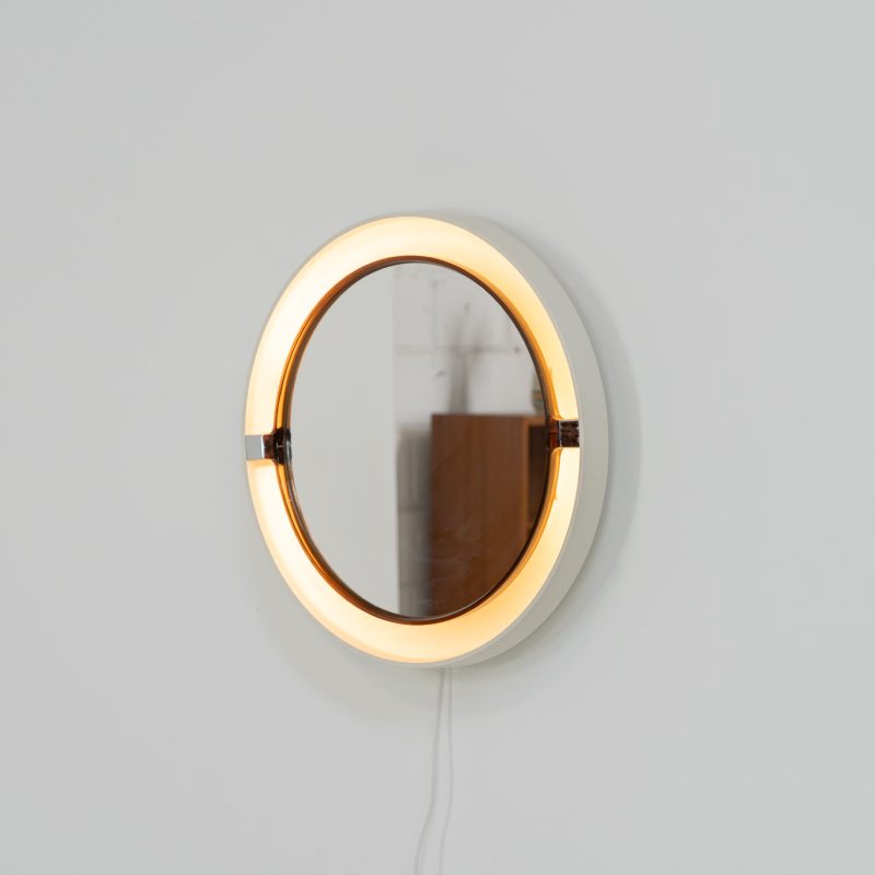 Model A41 backlit wall mirror by Allibert (Germany, 1970s). A striking space age design by Allibert, Germany. This Model A41 mirror is crafted from molded plastic and designed to hold four E14 bulbs inside, operated by a pull switch on the side. When illuminated, the translucent plastic frame softly diffuses the light, creating a warm halo effect around the mirror glass. The mirror can be tilted up or down, adding both functionality and a sculptural presence to the piece. Presented in good vintage condition with light, age-appropriate wear.
