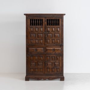 Stunning Spanish brutalist cabinet, 1950s. Striking and architectural, this 1950s Spanish cabinet is a powerful example of brutalist craftsmanship. Executed in solid wood, it features a deeply carved geometric façade with repetitive panel motifs that create a strong sense of rhythm and depth. The upper doors are accented with turned spindle details, adding contrast to the otherwise bold and graphic design. Original iron hardware, including hinges and latches, reinforces its rustic character. Behind the doors, the cabinet offers generous storage, making it as functional as it is sculptural. The warm patina of the wood enhances its presence, giving it a timeless, almost monolithic feel.