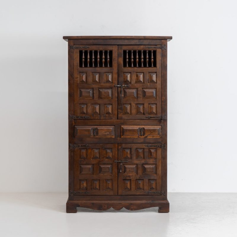 Stunning Spanish brutalist cabinet, 1950s. Striking and architectural, this 1950s Spanish cabinet is a powerful example of brutalist craftsmanship. Executed in solid wood, it features a deeply carved geometric façade with repetitive panel motifs that create a strong sense of rhythm and depth. The upper doors are accented with turned spindle details, adding contrast to the otherwise bold and graphic design. Original iron hardware, including hinges and latches, reinforces its rustic character. Behind the doors, the cabinet offers generous storage, making it as functional as it is sculptural. The warm patina of the wood enhances its presence, giving it a timeless, almost monolithic feel.
