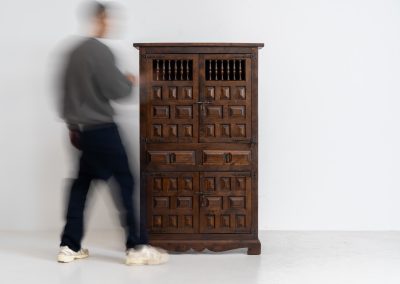 Stunning Spanish brutalist cabinet, 1950s. Striking and architectural, this 1950s Spanish cabinet is a powerful example of brutalist craftsmanship. Executed in solid wood, it features a deeply carved geometric façade with repetitive panel motifs that create a strong sense of rhythm and depth. The upper doors are accented with turned spindle details, adding contrast to the otherwise bold and graphic design. Original iron hardware, including hinges and latches, reinforces its rustic character. Behind the doors, the cabinet offers generous storage, making it as functional as it is sculptural. The warm patina of the wood enhances its presence, giving it a timeless, almost monolithic feel.