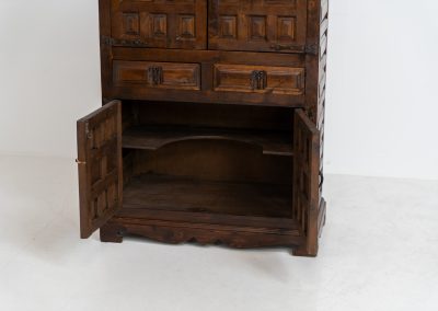 Stunning Spanish brutalist cabinet, 1950s. Striking and architectural, this 1950s Spanish cabinet is a powerful example of brutalist craftsmanship. Executed in solid wood, it features a deeply carved geometric façade with repetitive panel motifs that create a strong sense of rhythm and depth. The upper doors are accented with turned spindle details, adding contrast to the otherwise bold and graphic design. Original iron hardware, including hinges and latches, reinforces its rustic character. Behind the doors, the cabinet offers generous storage, making it as functional as it is sculptural. The warm patina of the wood enhances its presence, giving it a timeless, almost monolithic feel.