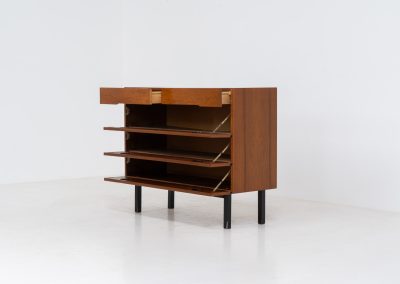 Teak shoe cabinet (Germany, 1960s). The cabinet features two small drawers and three shelves for shoe storage. It is made of teak and has a black lacquered frame with black inset handles that allow for proper ventilation. The cabinet is in good condition, with some visible signs of use.