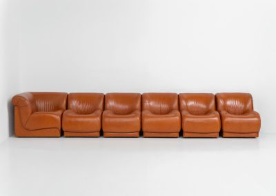 Modular sofa by Doimo Salotti (Italy, 1970s). Striking and bold modular seating system consisting of five identical elements and one corner piece, upholstered in their original cognac leather. Can be arranged as a sofa or used as individual lounge chairs. Low, sculptural design with generous padding, offering both strong visual impact and excellent comfort. The sofa is in very good condition with some age-related signs of use, as shown in the pictures. Most traces of use are discreet and not immediately visible. Marked by the maker.